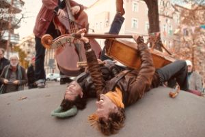 Zwei Männer liegen auf etwas gewölbtem. Einer hat ein Cello in der Hand. Eine weitere Person mit Cello steht neben ihnen.